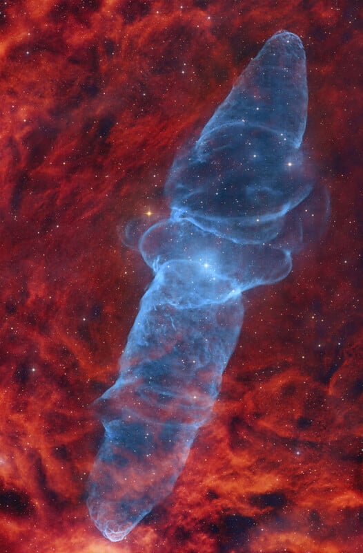 A large, blue, cloud-like nebula shaped like a cocoon floats in space, surrounded by a vibrant red and orange cosmic background filled with scattered stars.