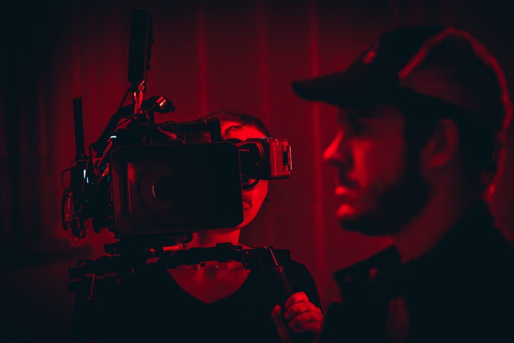 Image is a red and black photo of a camera pointing toward the profile of a man's face. He is wearing a baseball cap.