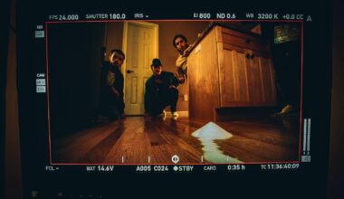 Image shows three people crouched on a wooden kitchen floor. One of the man is peering from behind a cabinet. The photo is framed in black with white camera markings.