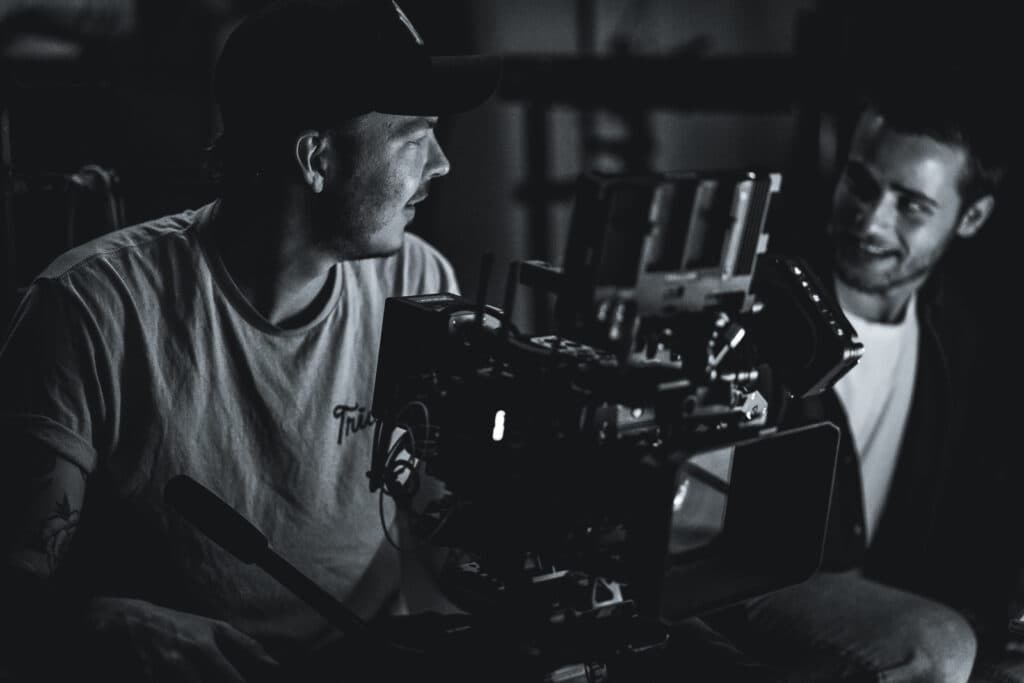 Image is a black and white photo of two people looking at each other with a movie camera in between them.