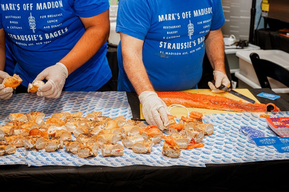 Two workers wearing latex gloves and blue Mark's Off Madison t-shirts arrange bagel samples on a tray