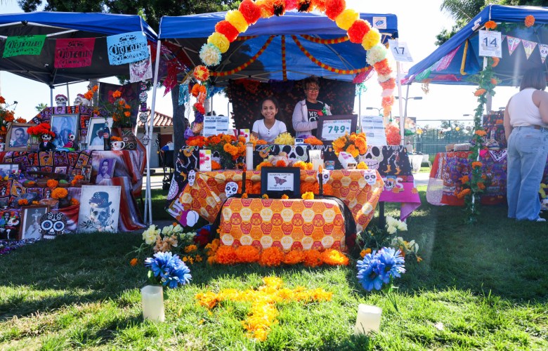 A vendor booth at a Dia de Los Muertos fest where a woman and a young girl stand behind an ofrenda decorated with orange cloth.