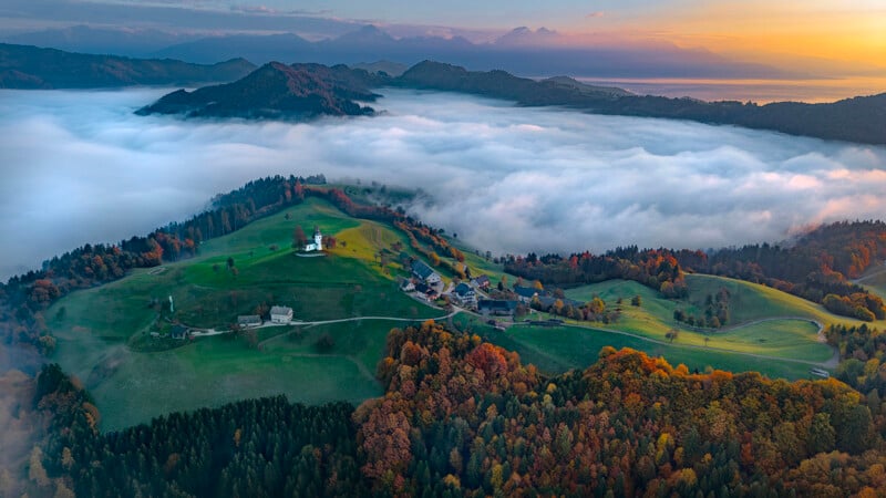 Aerial view of a green hillside village surrounded by autumn trees, with a church and houses, overlooking a valley covered in thick white fog and distant mountains at sunrise.