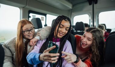 A front view of a group of three teenage girls on the minibus on the way to go on a hiking field trip. They are looking at one of the girls mobile phones and smiling and pointing at the screen.