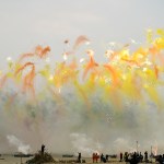 View of a fireworks display of mostly orange and yellow against a grey sky.
