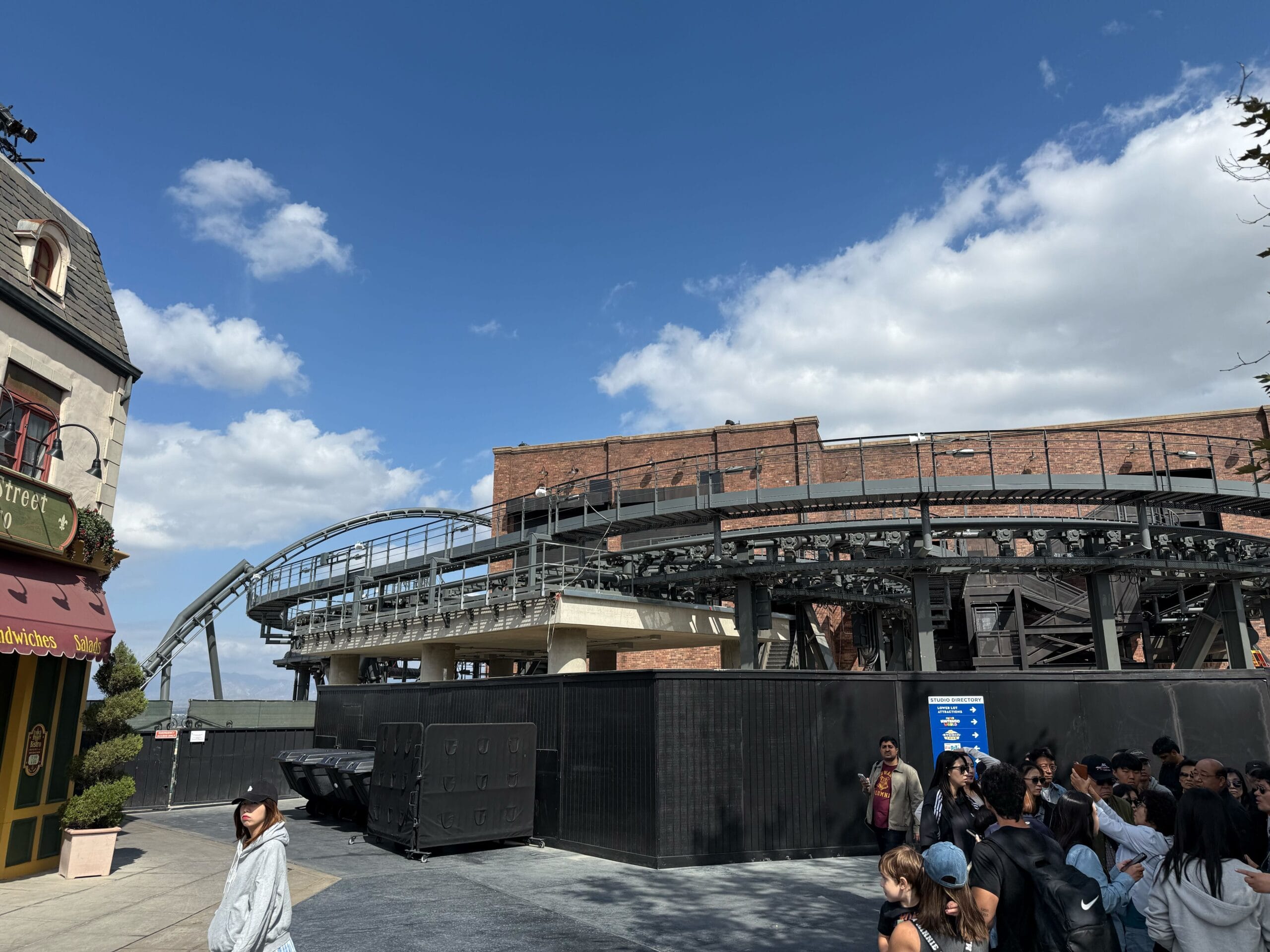 Guests gather by a roped-off roller coaster at a Disney park, beneath a bright blue sky dotted with clouds. No castle in view.
