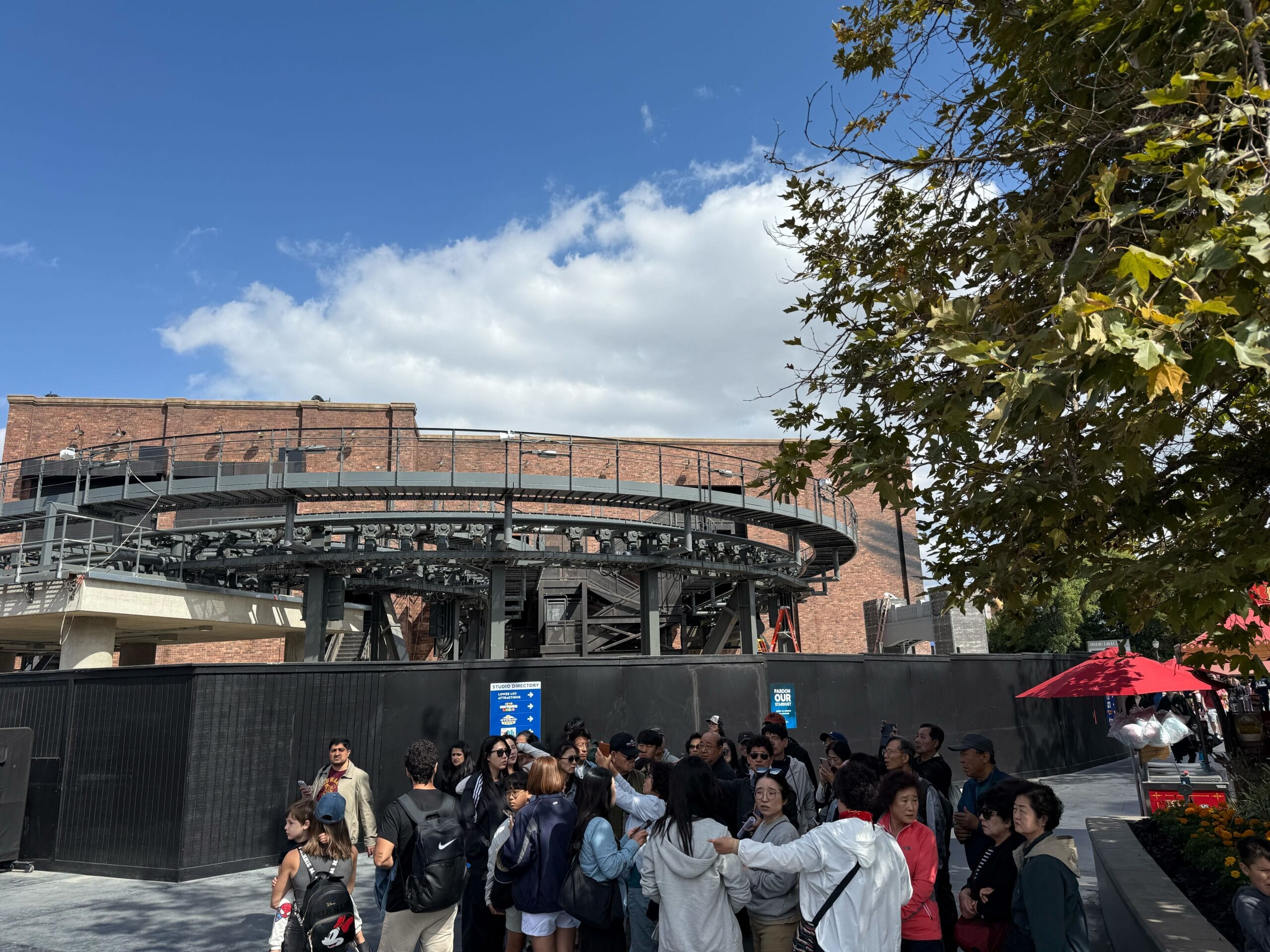 Guests gather near a fenced construction zone in a Disney park, with leafy trees and a sunny, cloud-dotted sky overhead.