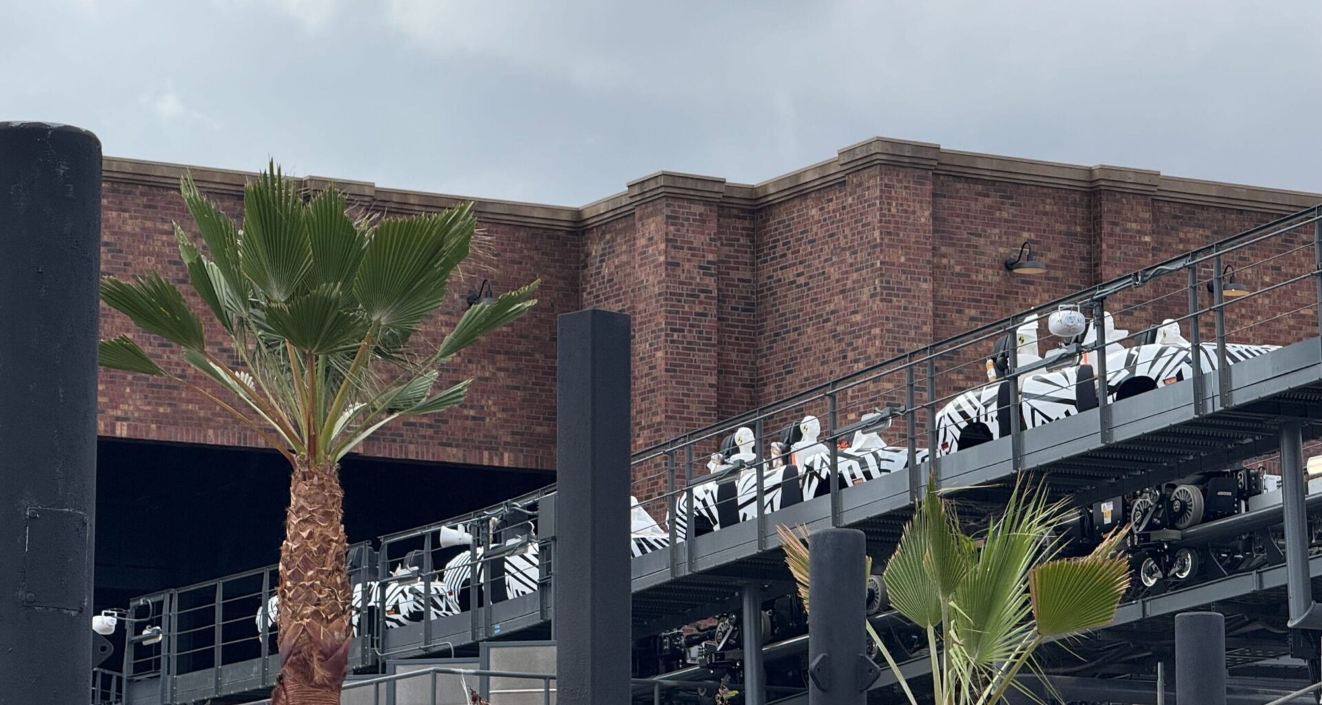 People in white-and-black uniforms ride a roller coaster near a brick building and palm trees, similar to Disney parks’ theming.