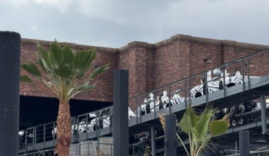 People in white-and-black uniforms ride a roller coaster near a brick building and palm trees, similar to Disney parks’ theming.