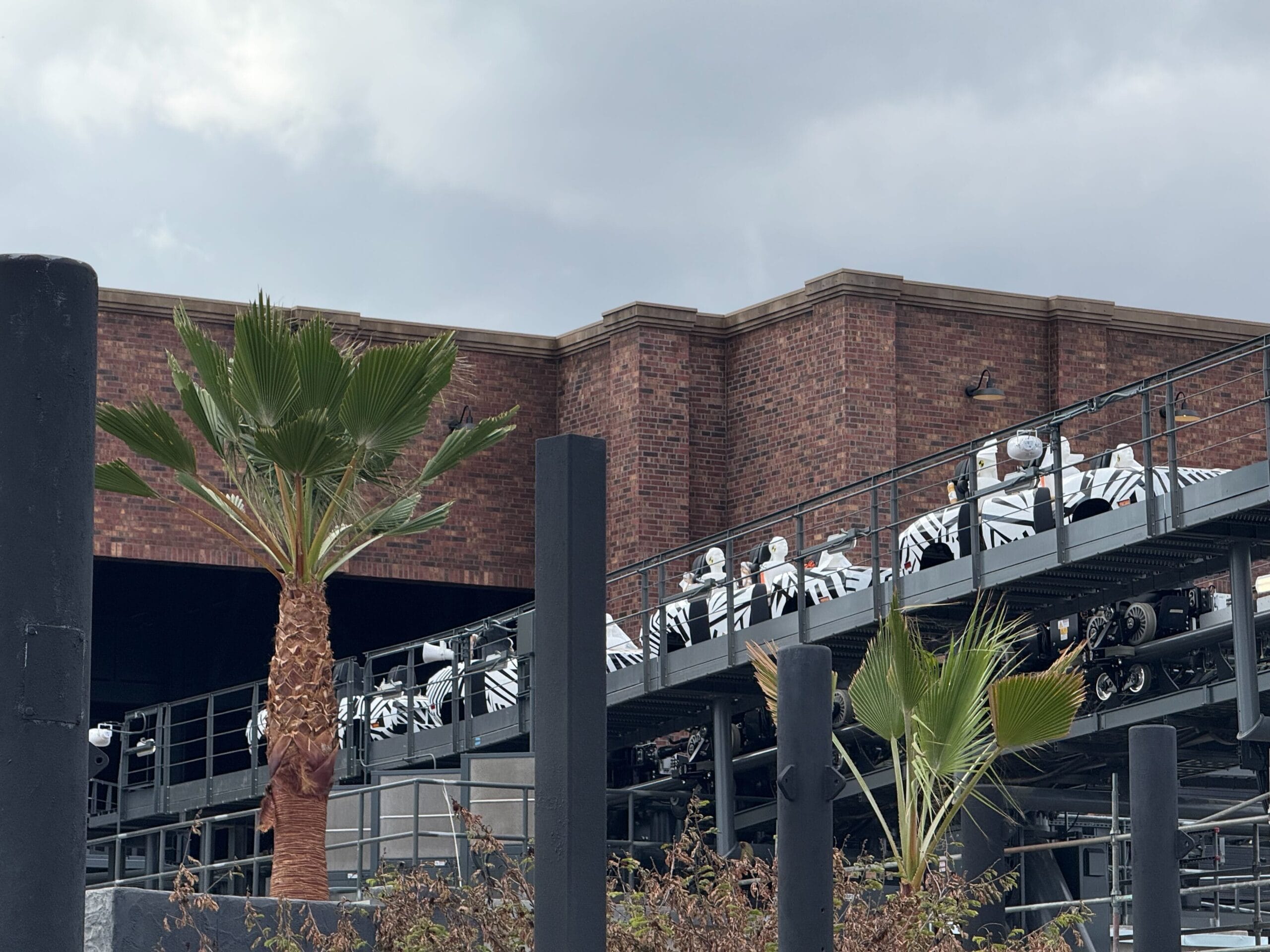 People in white-and-black uniforms ride a roller coaster near a brick building and palm trees, similar to Disney parks’ theming.