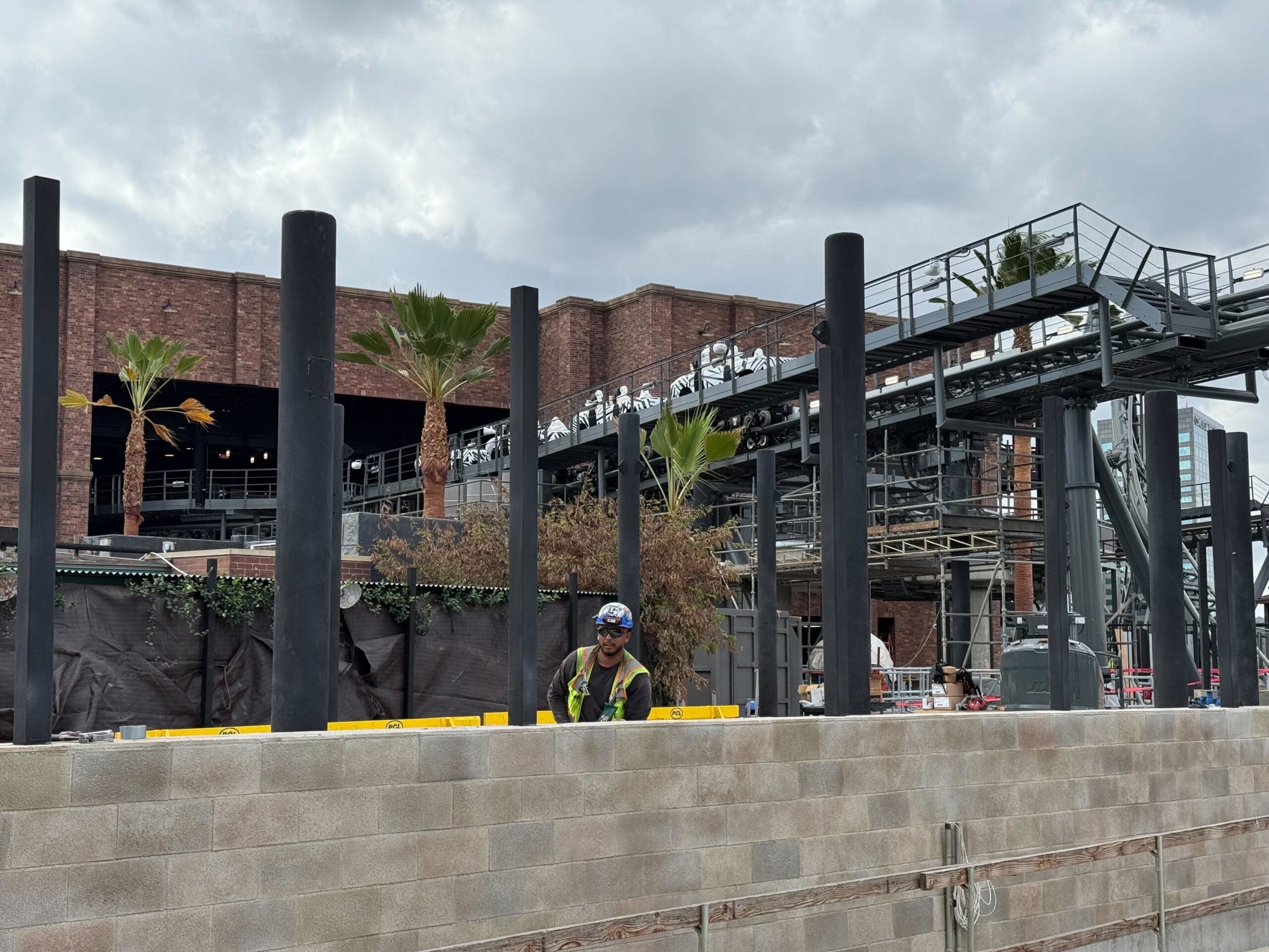 A construction worker stands by a brick building and scaffolding, with palm trees and stormy skies, not far from Disney park vibes.
