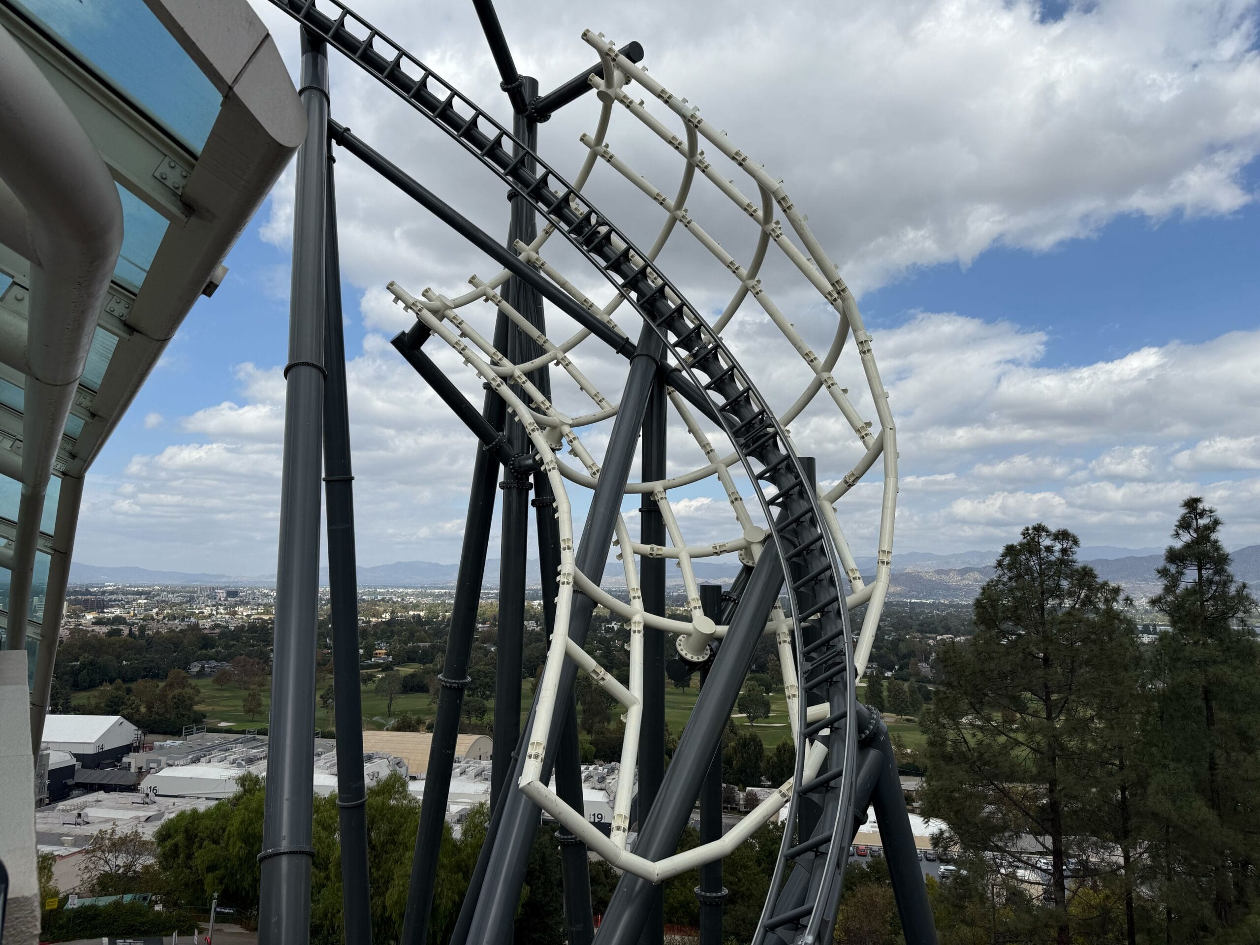 A steel roller coaster soars above treetops, city, and mountains—evoking thrills like Disney’s TRON Lightcycle, under cloudy skies.