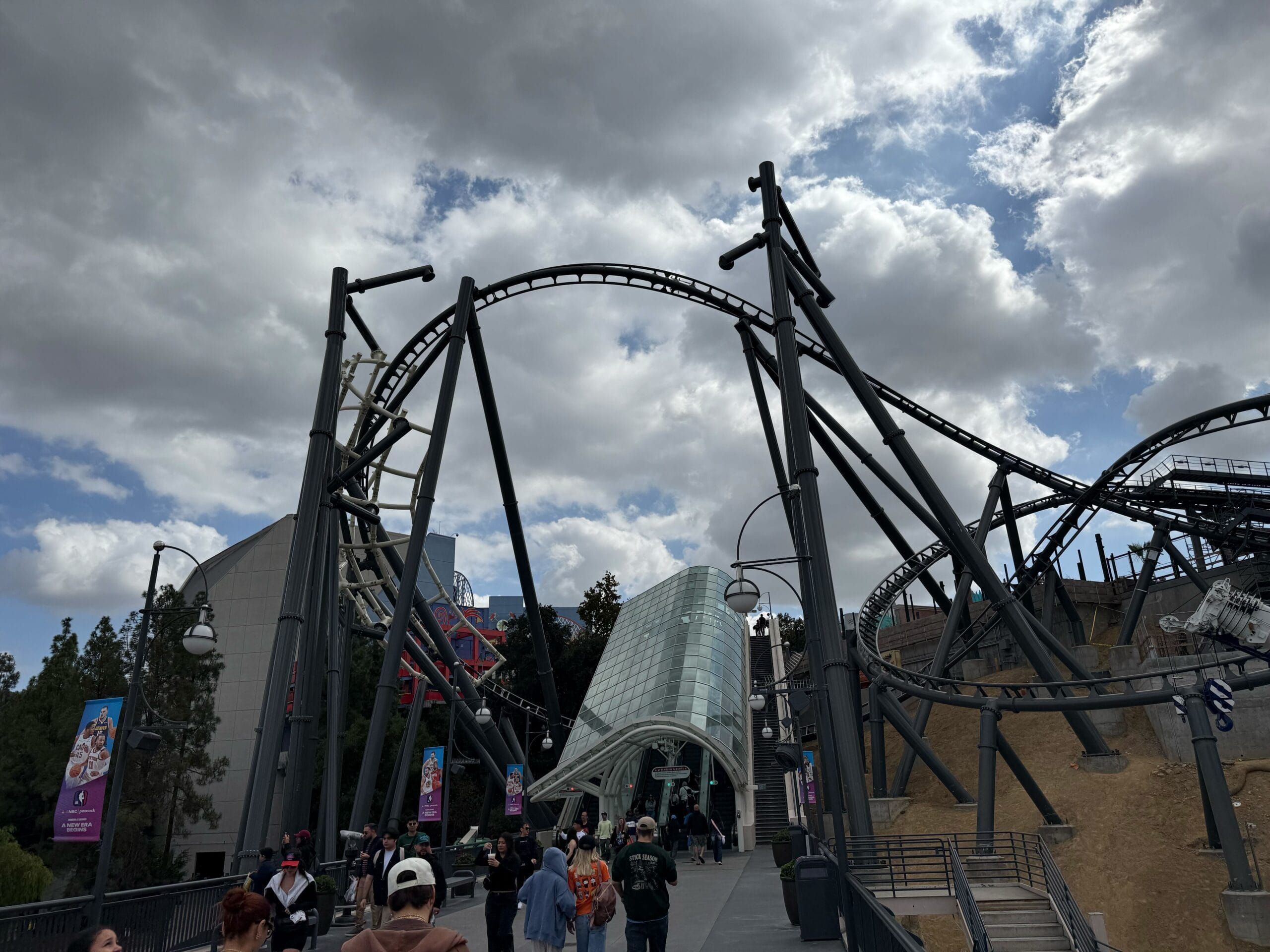 Guests stroll beneath a winding roller coaster track at a Disney park on a gray day, though no iconic castle is visible nearby.