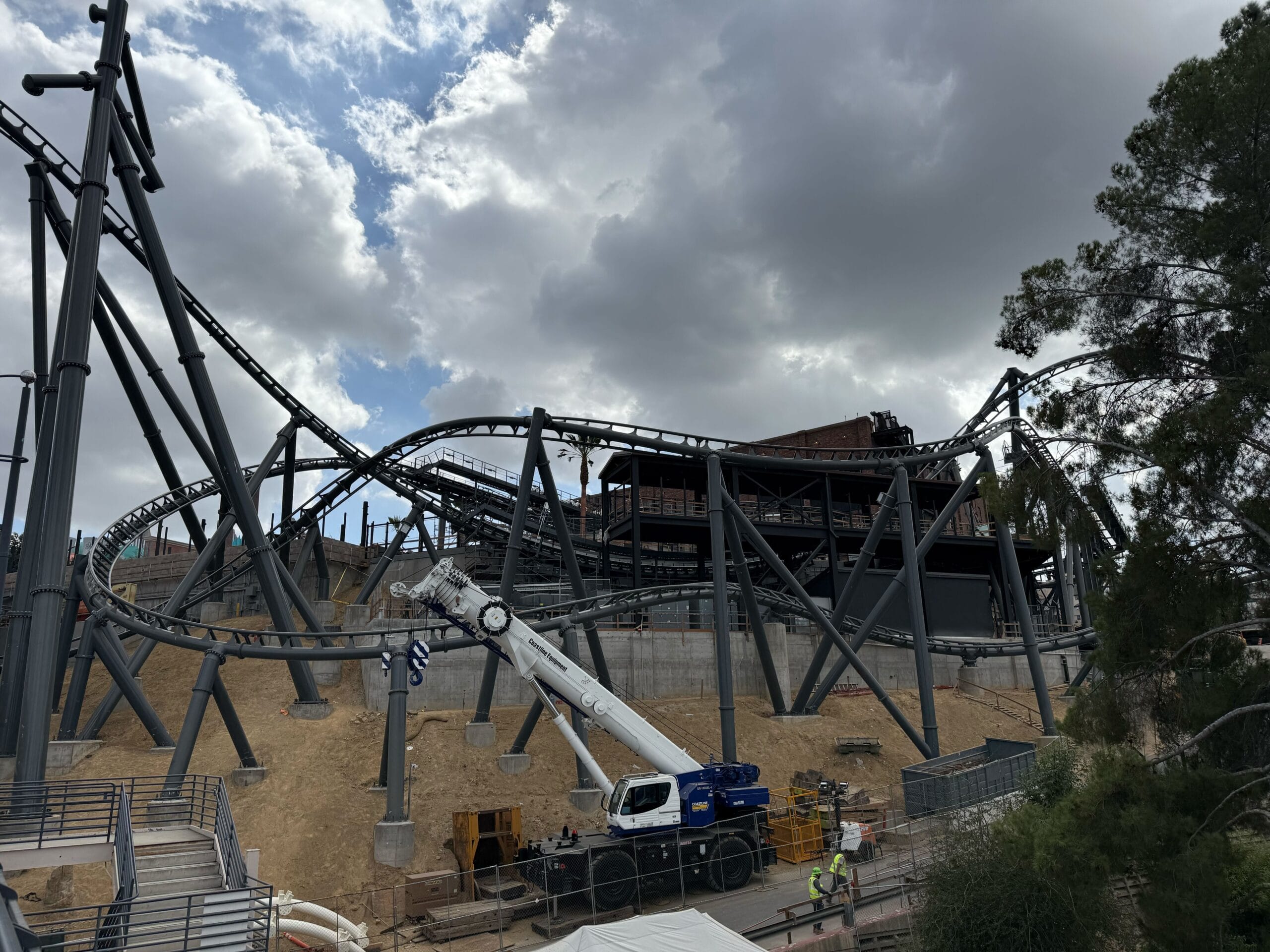 A roller coaster being built at a Disney park, crane and workers below a cloudy sky, not far from iconic Disney castle architecture.