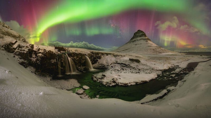 Snowy landscape with a mountain, waterfall, and river under vibrant green and pink Northern Lights illuminating the night sky.