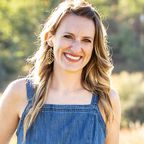 Caucasian woman with dark blonde hair and gold leaf earrings wearing denim tank top and smiling outside.