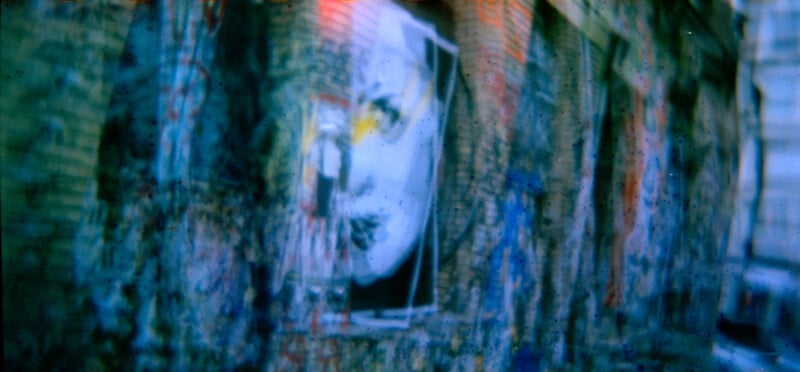 A blurred image of an old brick wall with graffiti and a large black-and-white poster of a face. The scene has a blue tint and an urban, gritty atmosphere.