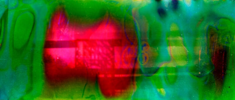 Abstract image with a vibrant mix of green, red, and blue hues, featuring a large, bright red area in the center surrounded by waves and patches of green, with faint geometric patterns and shadows throughout.