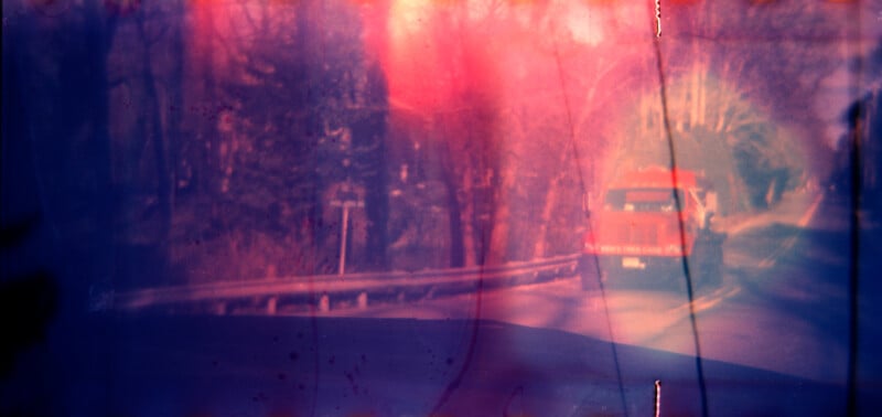 A red fire truck drives down a rural road lined with trees. The image has a blurry, dreamlike quality with streaks of pink, purple, and blue light effects overlaying the scene.