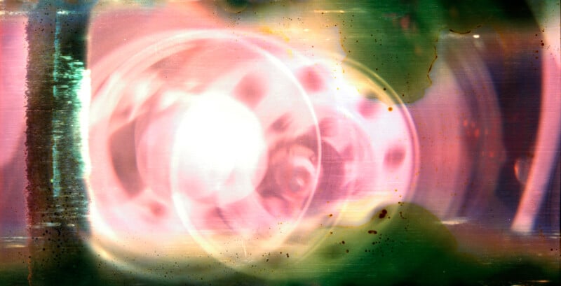 Abstract image with swirling pink, white, and green colors, featuring a bright circular light at the center, diffused shapes, and textures resembling film or a lens flare with scattered dark spots and streaks.
