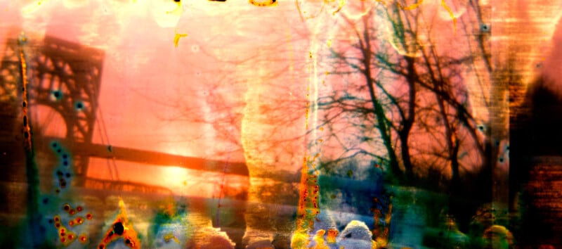 An abstract, colorful image featuring bare trees, a bridge, and a glowing sunset, with vibrant orange, yellow, and blue areas suggesting film damage or light leaks.