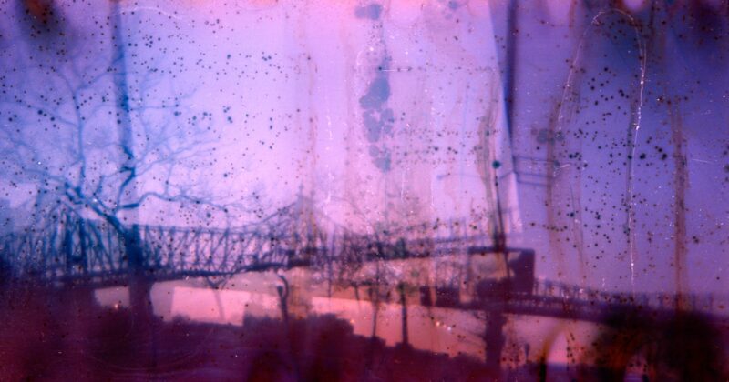 A silhouetted bridge and trees at sunset are visible through a hazy, textured layer of purple and pink colors, with spots and streaks creating an abstract, dreamy effect.