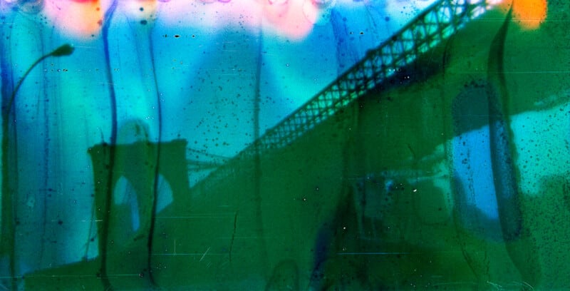 A blurry, abstract photo of the Brooklyn Bridge with colorful blue, green, and pink streaks and spots, creating an artistic, surreal effect over the iconic structure.