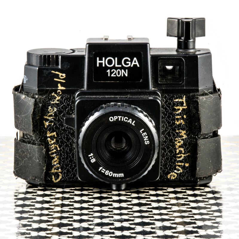 A black Holga 120N film camera with a 60mm optical lens sits on a checkered surface. The camera has worn black tape on both sides with handwritten yellow text.
