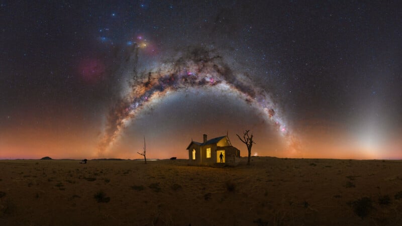 An abandoned house glows with warm light on a barren desert under a vivid night sky, where the Milky Way forms a bright, colorful arc overhead, framed by two leafless trees.