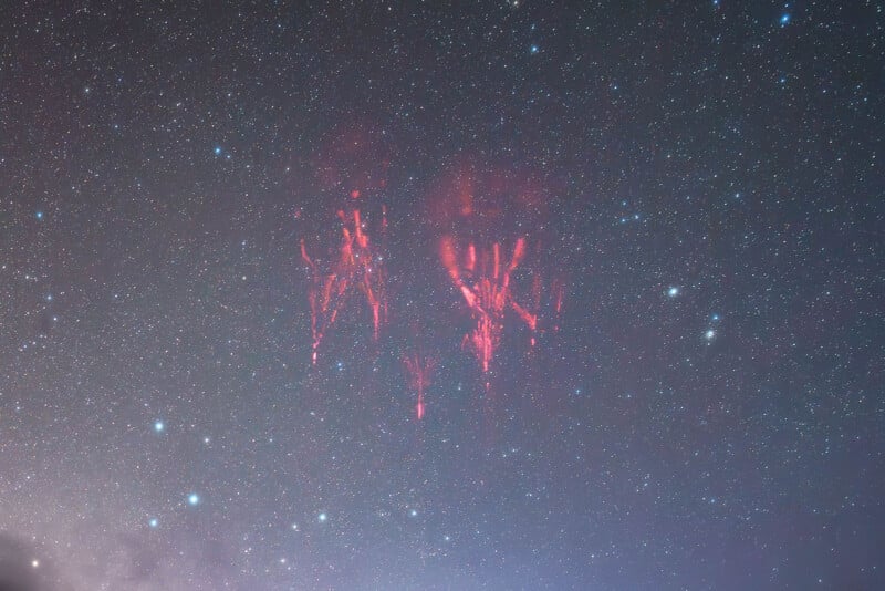 Red sprite lightning glows faintly in the night sky above a field of stars, creating streaks and tendrils of red light in the upper atmosphere amidst a clear, star-filled background.