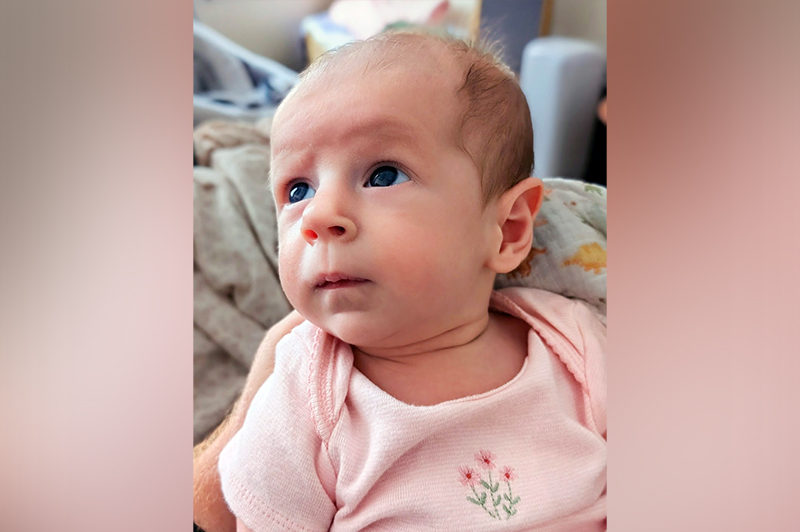 A baby in a pink onesie with floral embroidery gazes up and to the left while lying on a dinosaur-patterned blanket.