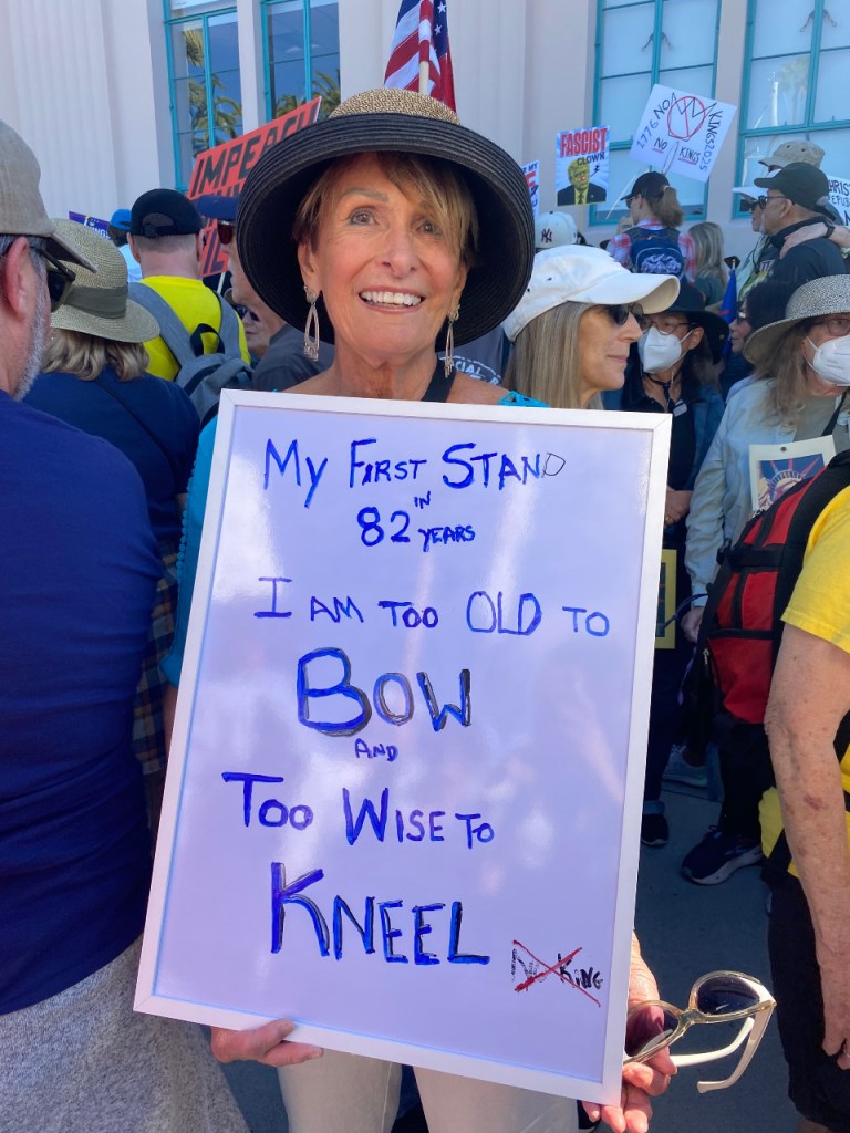 A woman in a hat holds a sign, proclaiming her "first stand in 82 years ... I am too old to bow and too wise to kneel.
