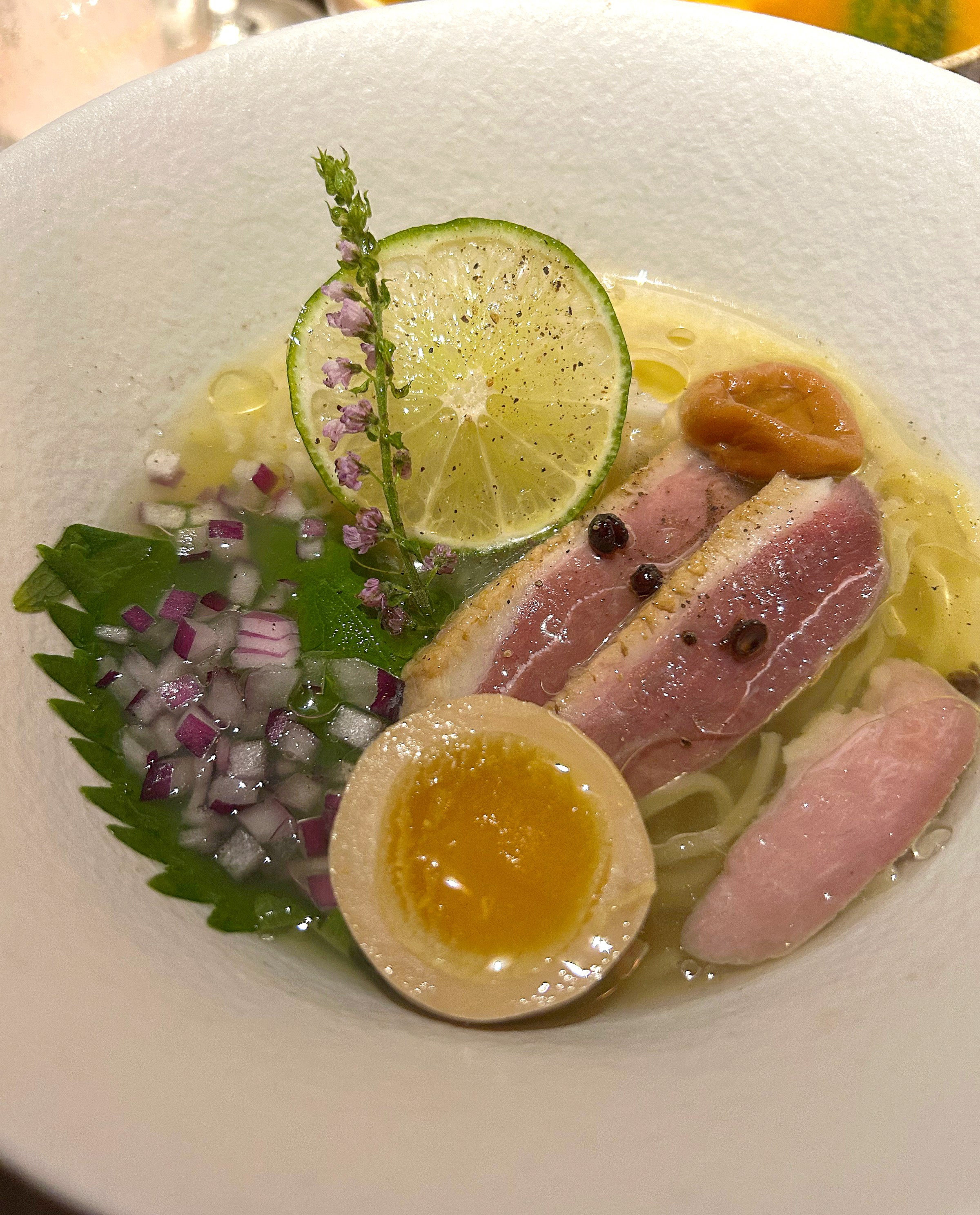 A bowl with sliced meat, diced onions, a slice of lime, a halved egg, and yellow broth.