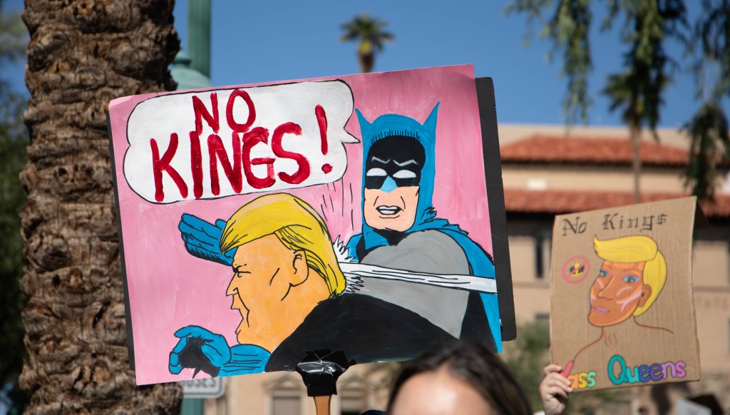 a protester holds a sign with batman slapping donald trump and saying "no kings"