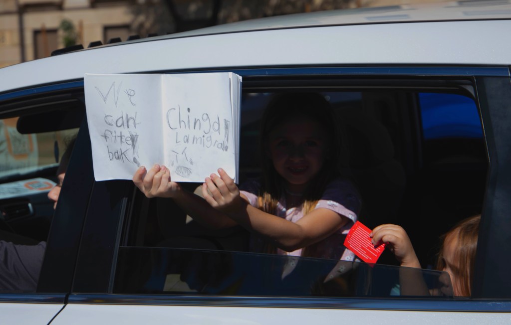 a girl holds a sign outside of a car that says "chinga la migra"