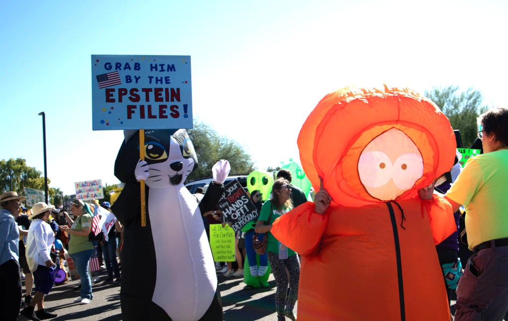 a protester in a cat costume holds a sign that says "grab him by the epstein files"
