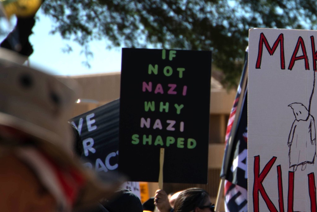 a protester holds a sign that says "if not nazi, why nazi shaped"