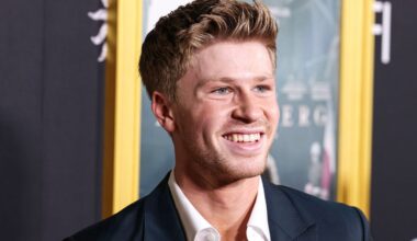 A young man with light brown hair, wearing a dark suit and white shirt, smiles while standing in front of a blurred movie poster at an event.