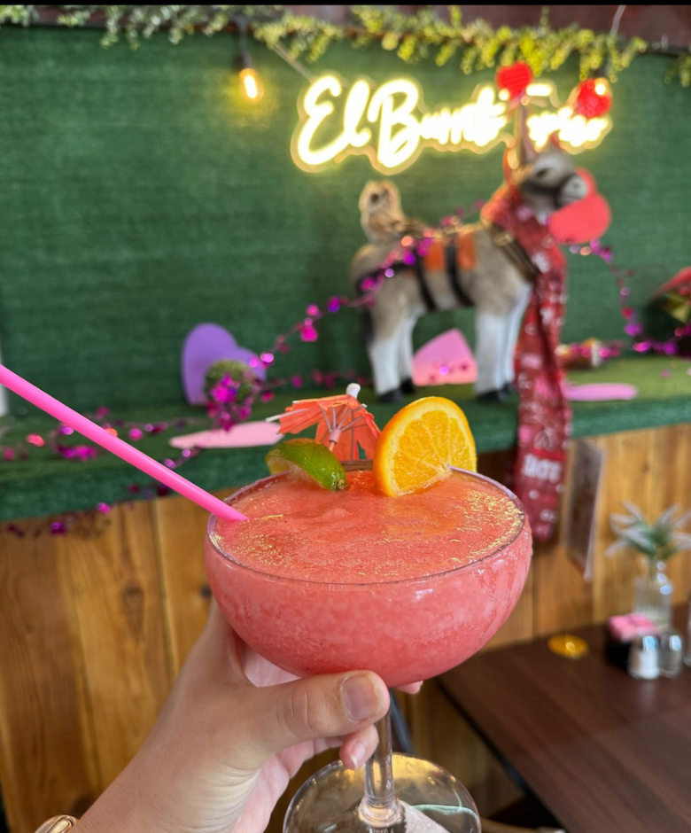 A hand holding a margarita inside the restaurant with a donkey behind it.