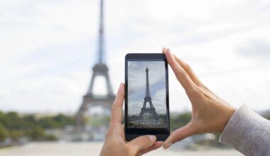 A person's hands hold a smartphone, taking a picture of the Eiffel Tower in Paris. The landmark is blurred in the background but clearly visible on the phone screen.
