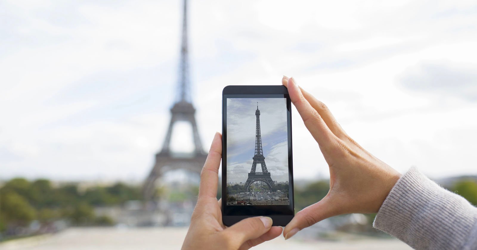 A person's hands hold a smartphone, taking a picture of the Eiffel Tower in Paris. The landmark is blurred in the background but clearly visible on the phone screen.