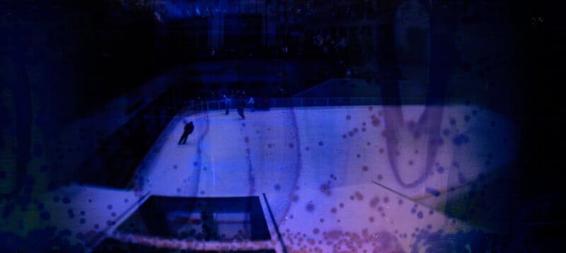An ice rink viewed from above, with a few people skating. The image is dark with a deep blue and purple overlay, featuring abstract spots and patterns across the scene.