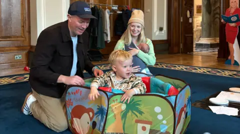 Mark McBride and his family are playing with a ball pit. Mark has a black jacket over a white t-shirt. He has a black cap on and brown trousers. His son is inside the ball pit, he has short blonde hair and a patterned coat on. His wife is behind the ball pit, she is wearing a green jumper and brown hat. She has their daughter in her arms. The background is a busy room with blue carpet and wooden walls. 