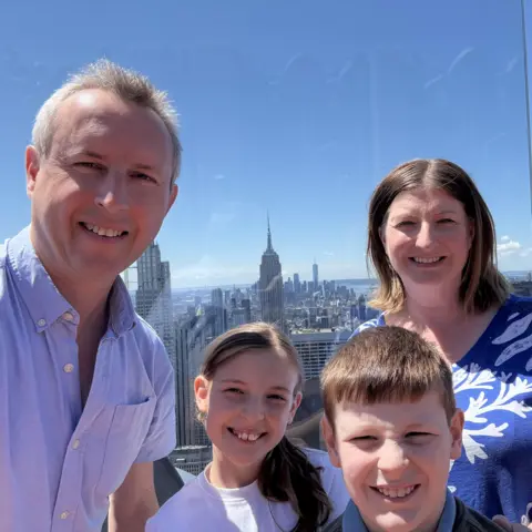 David Harris David Harris and his family take a selfie while posing on top of the Rockefeller Center in New York. The New York skyline and its skyscrapers can be seen in the background.