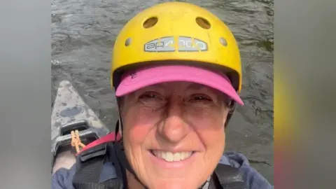 Justine Evans Wildlife filmmaker Justine Evans takes a selfie while canoeing on the river Wye.  She is sat in a black kayak, wearing a dark blue hoodie, a red life jacket and a pink baseball cap.  