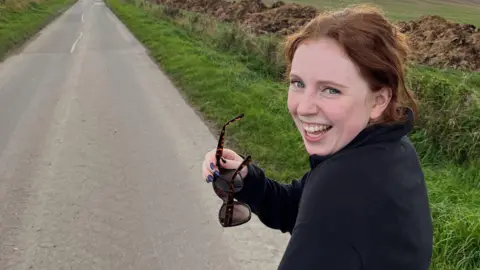 Liv Heeney A woman with red hair and wearing a black jacket smiles at the camera over her shoulder. She is holding a pair of brown and black sunglasses in her hand. She is walking on a rural country road.