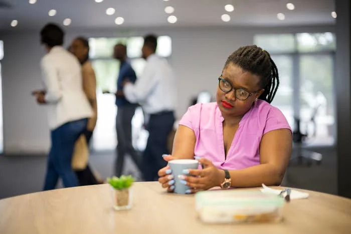Person holding a mug, seated at a table in an office setting, appearing thoughtful. Other people are walking in the background