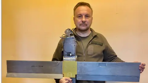Serhiy Beskrestnov Serhiy Beskrestnov is a middle-aged man wearing a khaki uniform. He is holding a drone with a wingspan of just under a metre and a half. It looks quite roughly put together - crudely constructed. Serhiy is looking down a barrel of the camera. 