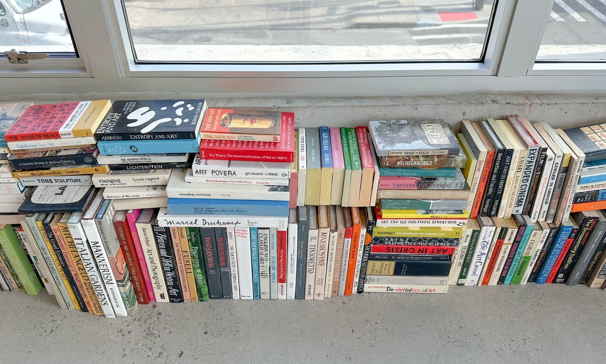 a collection of wooden and painted book sculptures by Conrad Bakker stacked on the floor beneath a window