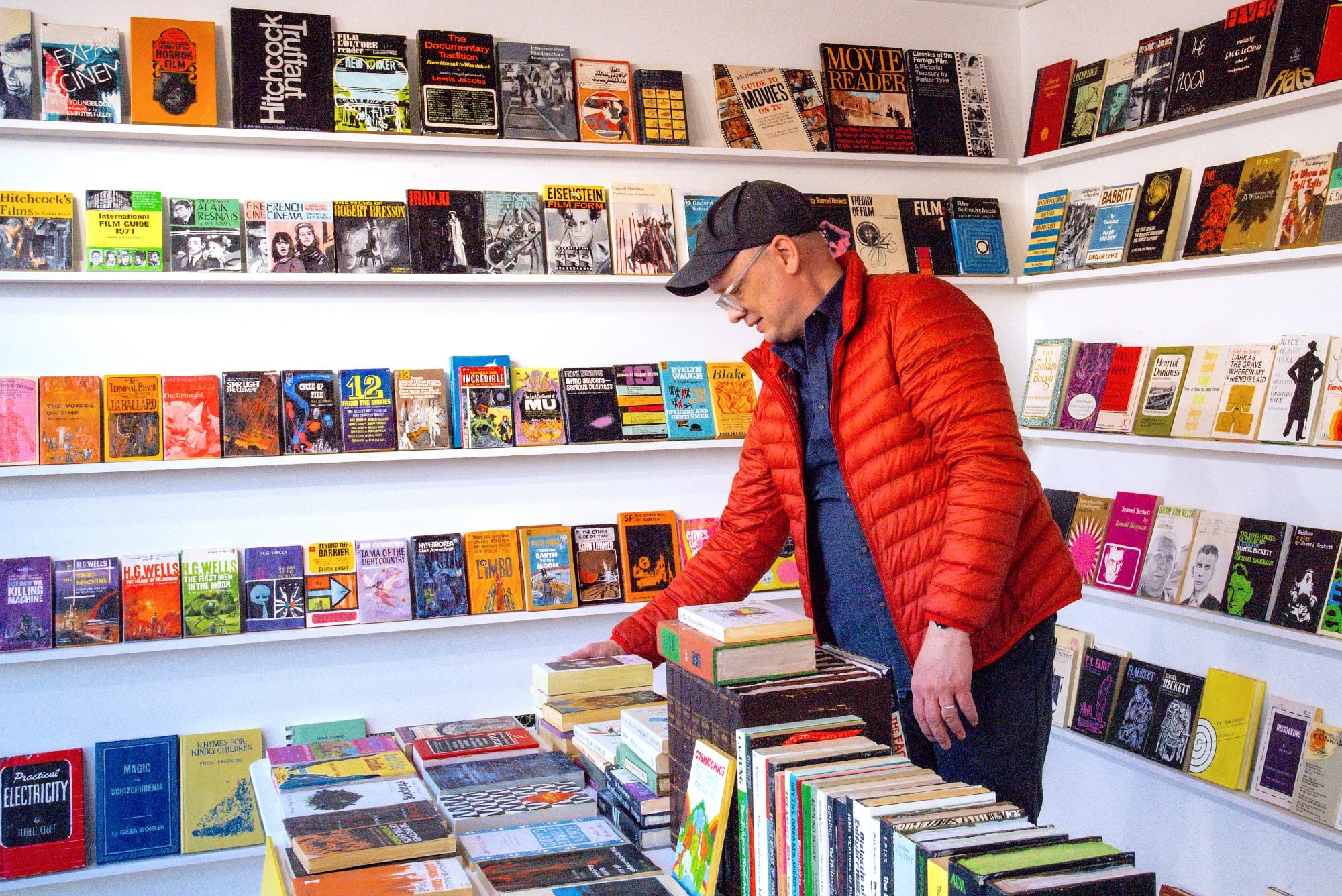 a collection of wooden and painted book sculptures by Conrad Bakker in boxes on a table and lining a wall. the artist stands in the center in a hat and puffy orange jacket